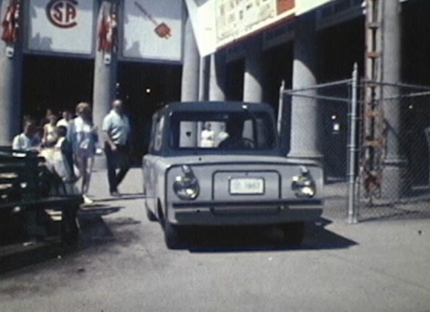 A boxy vehicle being driven through an outdoor exhibition space, with passers-by watching in the background.