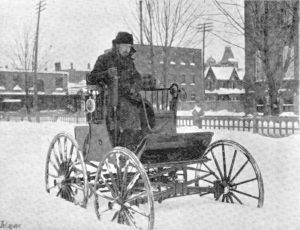 Black and white photograph of a moustached man driving a high-seated electric car through deep snow.