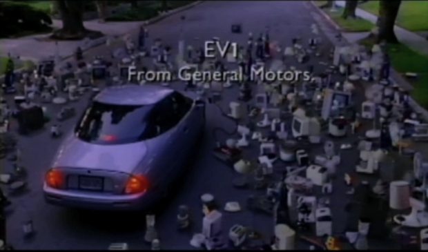 Rear shot of an electric car parked in the middle of the road, surrounded by a crowd of hundreds of electric appliances. Text reads “EV1 / From General Motors.”