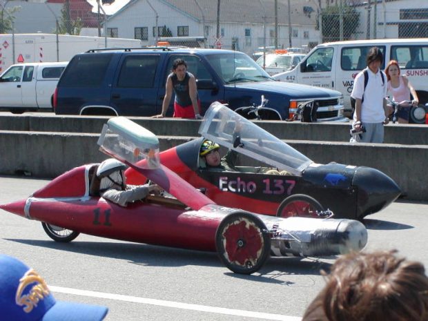 Colour photograph of two teenagers parked in streamlined homemade electric race-cars at the edge of a race track.