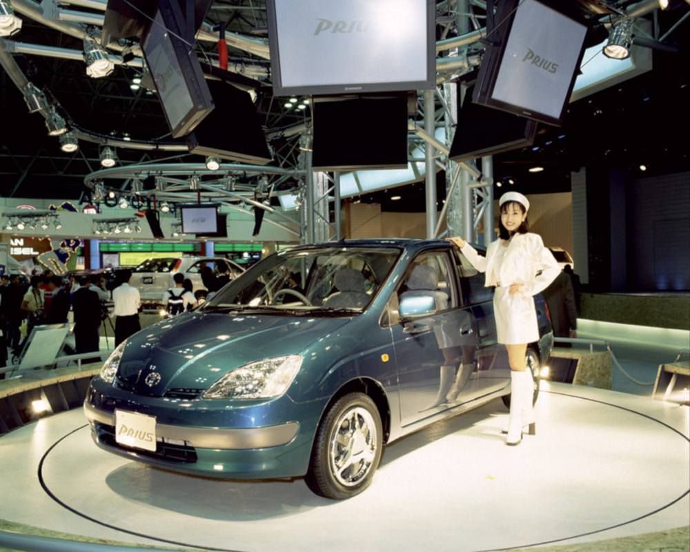 Photo couleur d'un mannequin qui pose à côté d'une voiture électrique sur un podium d'exposition lors d'un salon de l'automobile. Des écrans placés au-dessus du podium affichent: Prius.
