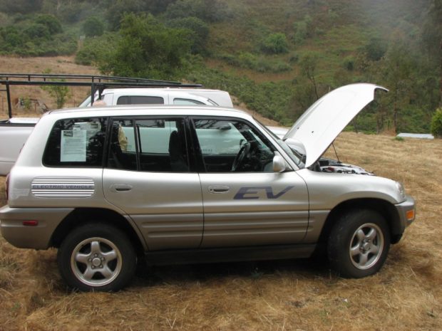 Colour photograph of a compact SUV with its hood open parked on a grassy hillside. Signage on the door reads “EV”