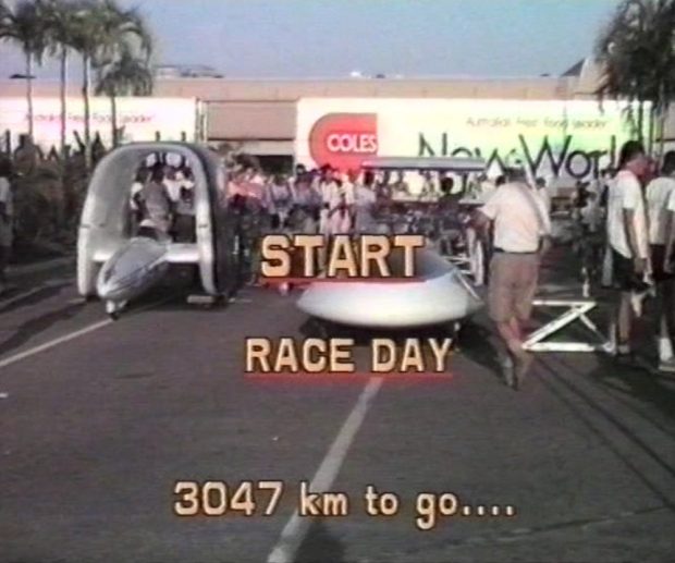 Several unusually-shaped vehicles are parked at the start line of a crowded racetrack, surrounded by crew and technicians. Overlaid text reads “START / RACE DAY / 3047 km to go…”