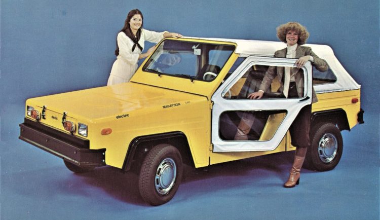 Two smiling women stand in the doors of a convertible two-seater electric truck.