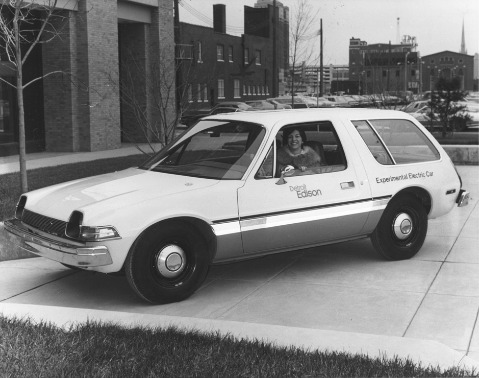 Black and white photograph of a smiling woman leaning out the driver’s seat of a compact station wagon. Writing on the side of the vehicle reads “Detroit Edison / Experimental Electric Car.”