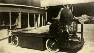Black and white photograph of a flat-bed industrial vehicle in a lumber yard, with stacks of wood in sheds in the background.