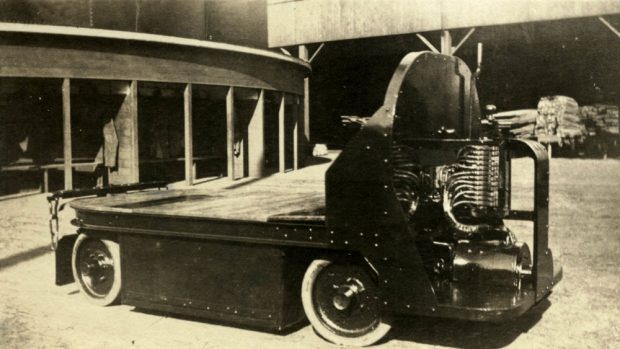 Black and white photograph of a flat-bed industrial vehicle in a lumber yard, with stacks of wood in sheds in the background.