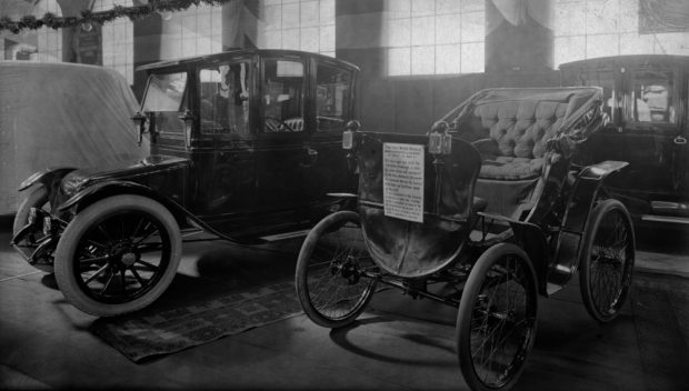 Two electric cars displayed next to each other in an exhibit space. The smaller is fitted with a sign reading THE FIRST MOTOR VEHICLE.