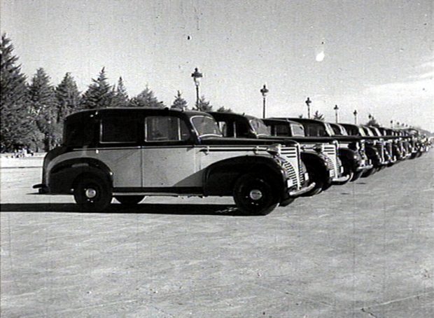 Black and white image of a line of automobiles parked in an empty lot, with pine trees and decorative street lamps in the background.