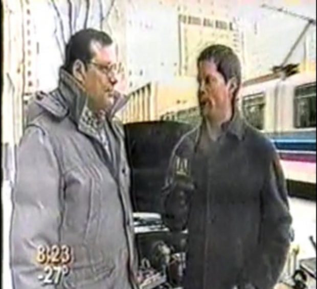 Image of two men in heavy winter gear standing in front of a car with an open hood on a city street, with a streetcar passing in the background. The man on the right is holding a microphone. Text at the bottom left of screen reads “8:23 -28°”.