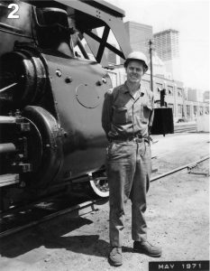 Black and white archival photograph of a smiling man stands wearing a hard hat and overalls in front of a steam locomotive, which is only partially visible, on the left side.