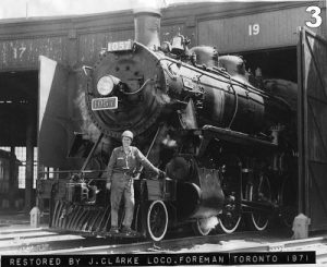 Black and white archival photograph of a smiling man in a hard hat standing on the front of a large steam locomotive. The locomotive is emerging from a roundhouse.