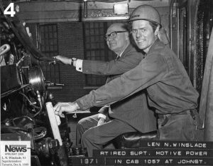Photo d’archive en noir et blanc de deux hommes dans la cabine d’une locomotive. Ils prennent la pose pour l’appareil photo. L’homme au premier plan porte une combinaison et un casque de sécurité et l’homme au second plan porte un costume.
