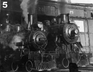Black and white archival photograph of two steam locomotives moving out of a roundhouse. There is steam billowing from their stacks and the stall doors are marked 27 and 28 above them.