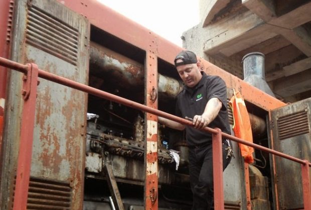 Colour photograph of a man standing on the platform of a locomotive with engine doors open. He is looking down and has a tool in his hand.