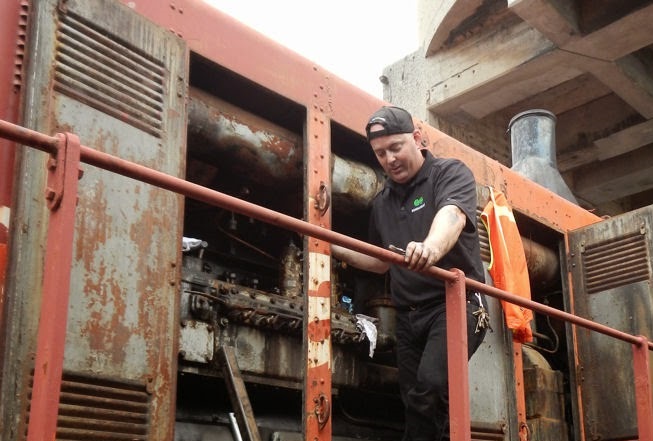 Colour photograph of a man standing on the platform of a locomotive with engine doors open. He is looking down and has a tool in his hand.