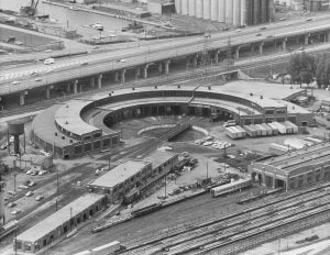 Black and white vintage aerial photograph showing a roundhouse building and railway tracks. There is a highway in the background and two rectangular buildings in the foreground.