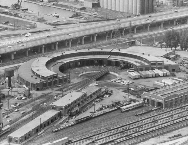 Black and white vintage aerial photograph showing a roundhouse building and railway tracks. There is a highway in the background and two rectangular buildings in the foreground.