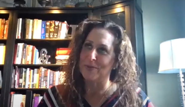 Colour screen capture of a woman smiling and looking up. Head and shoulders are visible, with shelves of books in the background.