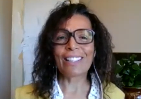 Colour screen capture of a woman with glasses and earphones smiling at the camera. Head and shoulders are visible.