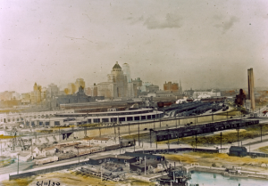 Colour painting of the cityscape shows many small buildings in the foreground. There is a roundhouse and railway tracks in the centre of the painting. The background shows the cityscape with taller skyscrapers including the Royal York Hotel.