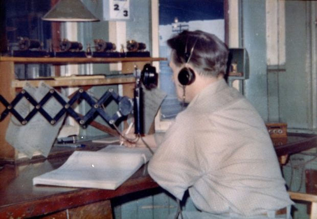 Colour vintage photograph of a man is sitting at a desk and writing in a large book. He is wearing a headset and a vintage phone is stretched towards him.