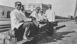 Black and white archival photograph of 5 Black men sitting on a railway track. They are dressed in white railway uniforms.