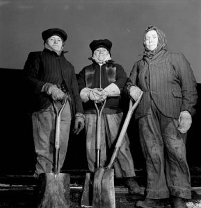 Black and white archival photograph of three women posing with shovels. They are dressed in coats and hats, scarves and work gloves.
