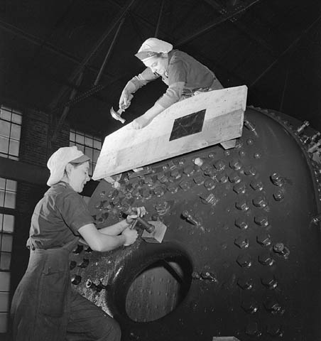 Black and white archival photograph of two women working on a large locomotive boiler. One woman is on top of the boiler and hammering wood that is affixed to the boiler. A second woman is on the ground working on the boiler.