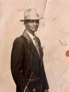 Sepia-toned black and white archival photograph of a man from the waist up. He is wearing a hat and suit and smiling slightly at the camera.