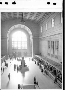 Black and white archival photograph of the interior of a large railway station. The photograph is taken from above and shows many ticket counters on the right hand side. There are several people walking around the station.