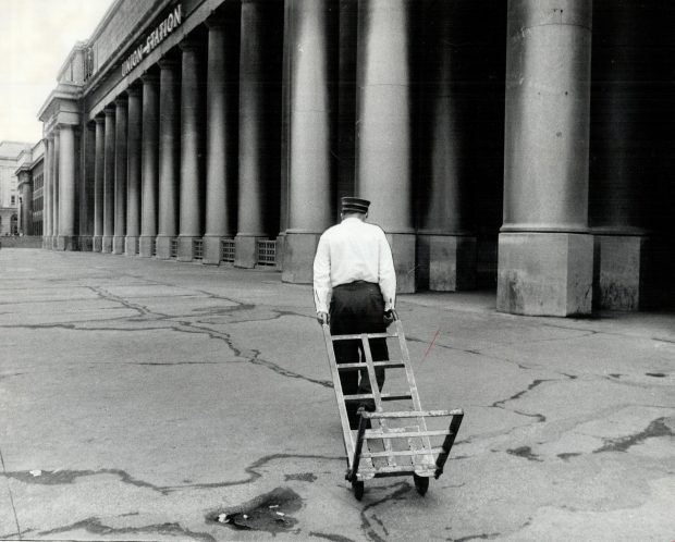 Black and white archival photograph of a man in railway uniform pulling an empty baggage cart in front of Toronto’s Union Station. He is walking away from the camera.