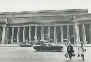 Black and white archival photograph of the front of a large railway station. There are vintage cars driving on the street in front of the station and two women carrying shopping bags in the foreground.