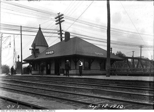 Black and white vintage photograph of a small train station with railway tracks in the foreground. A couple people are seen in front of the station. “DON” is written twice on the roof of the station. On the photograph “Sept. 12. 1910” is written in the bottom right corner.