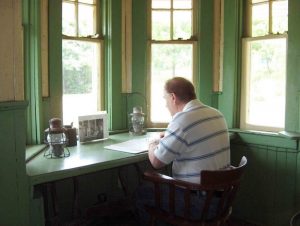 Colour photograph of a man is sitting at a desk facing away from us. There is an open binder and 2 railway lanterns on the desk. There are 4 windows facing the desk and it is a sunny day.