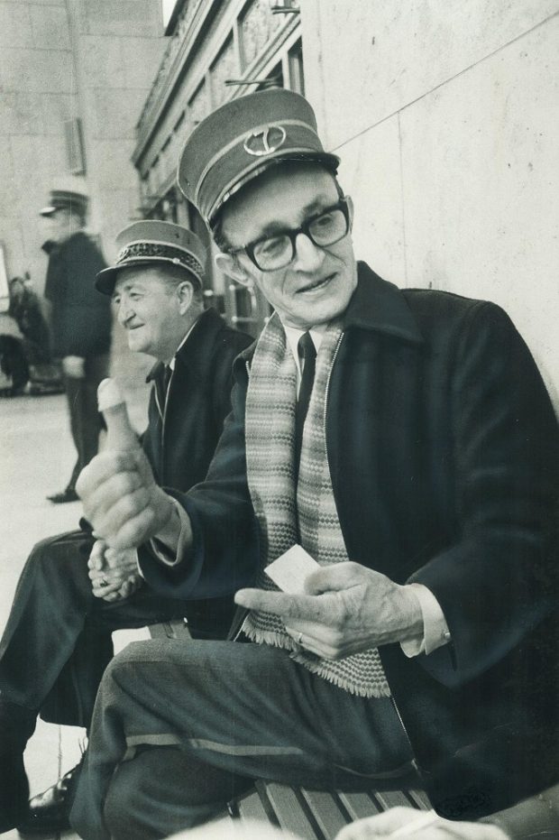 Black and white archival photograph of two men sitting on a bench. The men are wearing railway uniforms including hats. The man in the foreground is speaking and gesturing with his hands. The man in the background is looking away from the camera.