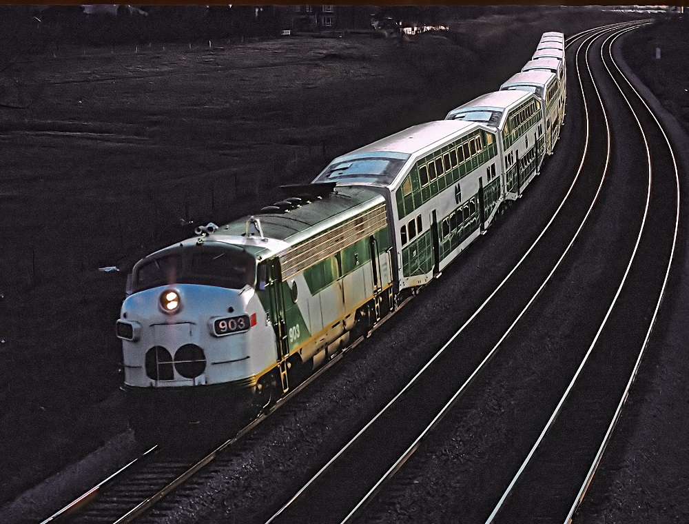 Colour photograph of a GO Transit locomotive and seven rail cars travelling down a railway track. There are two empty tracks next to the train.