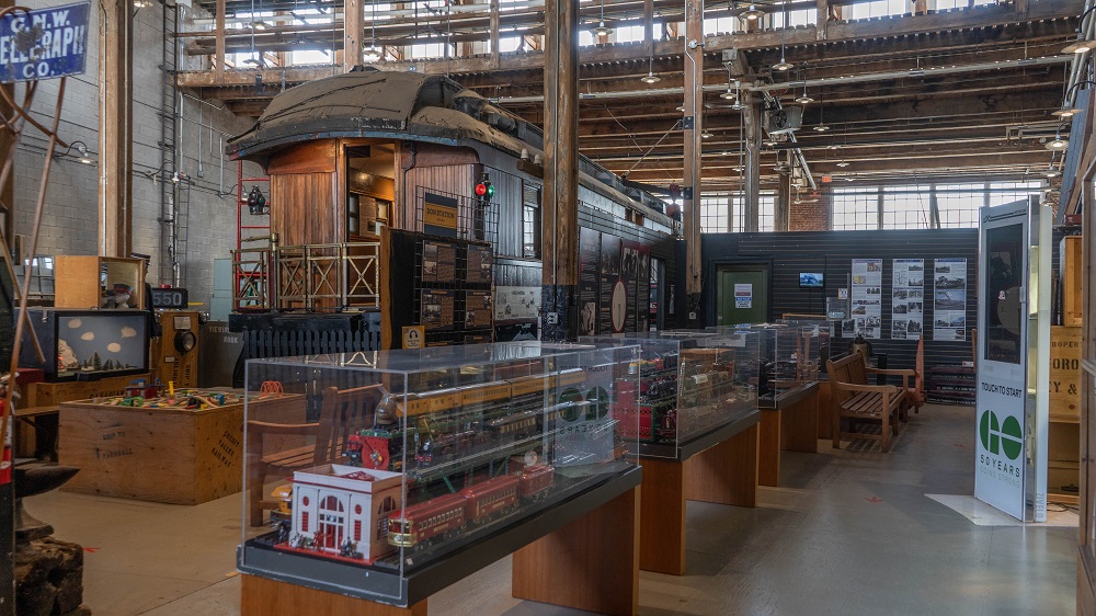 Colour photograph of the interior of a museum. There are several exhibit cases of model trains in the foreground and a railway car in the background.