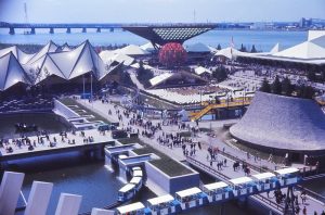 Colour archival photograph of Expo 67. The photograph is taken from above and shows four short buildings with unusual shapes. There are many people walking on the roads between the buildings.