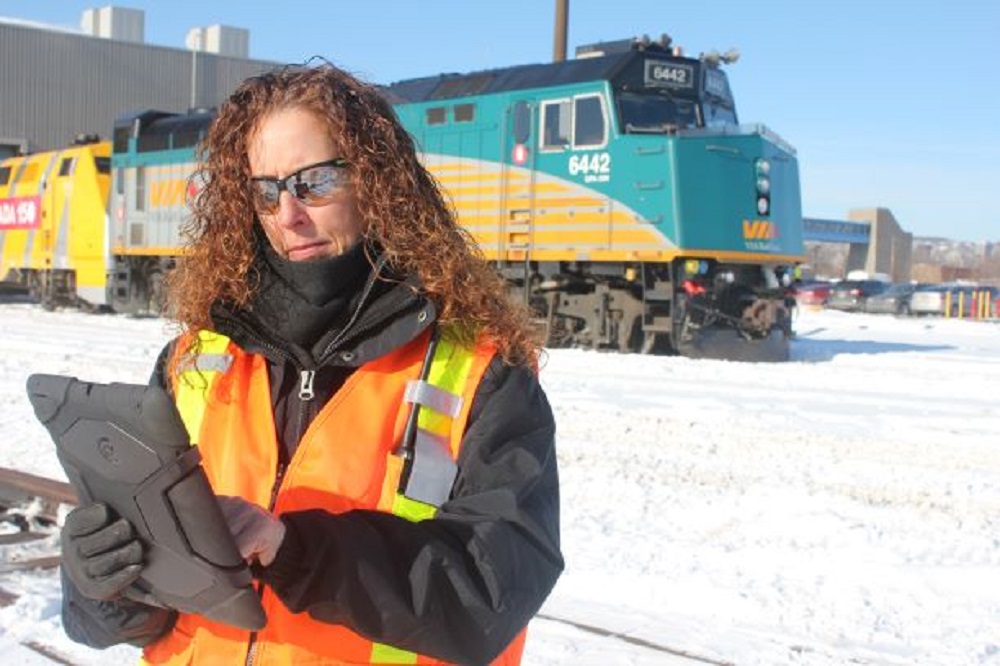 Modern colour photograph of a woman in a high visibility vest working on a tablet. There is a large green locomotive in the background.