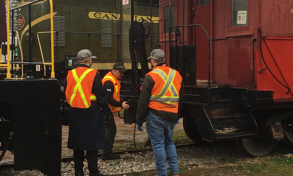 Colour photograph of three men in high-visibility vests standing between a locomotive and caboose. Two of the men are facing away from the camera. The man in the centre is faced toward the camera and is pushing a coupler on the caboose.