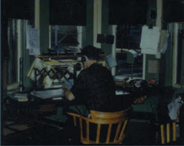 A vintage colour photograph of a man working at a desk. He is facing away from us. He is wearing a headset and a vintage phone is pulled towards him. The desk has many papers on it.
