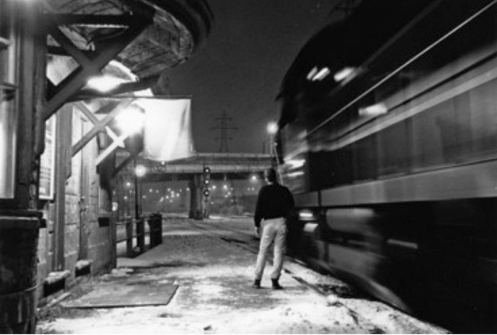 Black and white photograph of a man stands on a railway platform while a train is driving by on the right side. He is holding a large stick up to the train. Part of the train station can be seen on the left side.