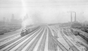 Black and white vintage photograph showing railway tracks. There is a steam locomotive on the left side with steam billowing upwards. The background is obscured by fog.