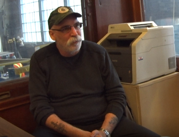 A colour still image captured of a video recording. A man is looking off camera with his hands folded in his lap.