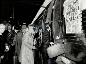 Photographie d’archive en noir et blanc d’un groupe de personnes montant dans un wagon de train de passagers. Une affiche sur le wagon indique : « BIENVENUE À BORD DU TRAIN INAUGURAL TR. 64 MAPLE LEAF TORONTO À NEW YORK 26 AVRIL 1981. CHOO-CHOO BOB. »