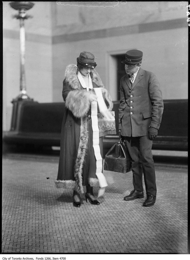 Black and white archival photograph of a lavishly dressed woman flipping through a long train ticket while a uniformed man also looks at the ticket.