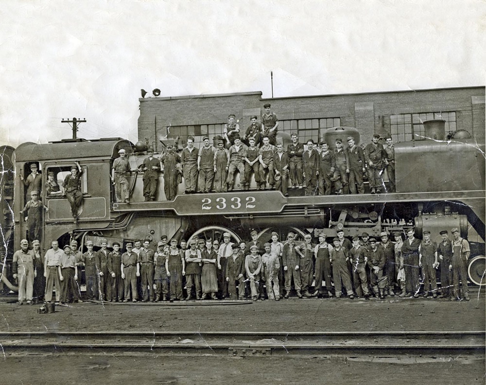 Black and white archival photograph of approximately 75 people posed in front of and on top of a large steam locomotive on railway tracks. The locomotive number “2332” is visible in the centre of the locomotive. There is another set of empty railway tracks in foreground and a short rectangular building in the background.