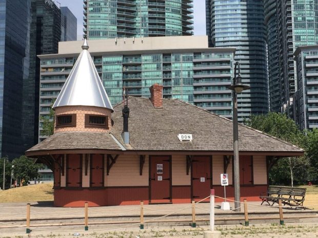 A modern colour photograph of a small train station with ropes and miniature train tracks in the foreground. “DON” is write on the roof of the station and there is a small sign that says “GIFT SHOP” in front of the station.