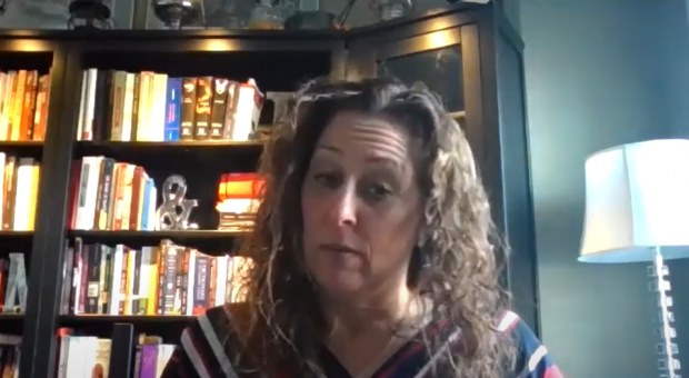 Colour screen capture of a woman looking down. Head and shoulders are visible, with shelves of books in the background.